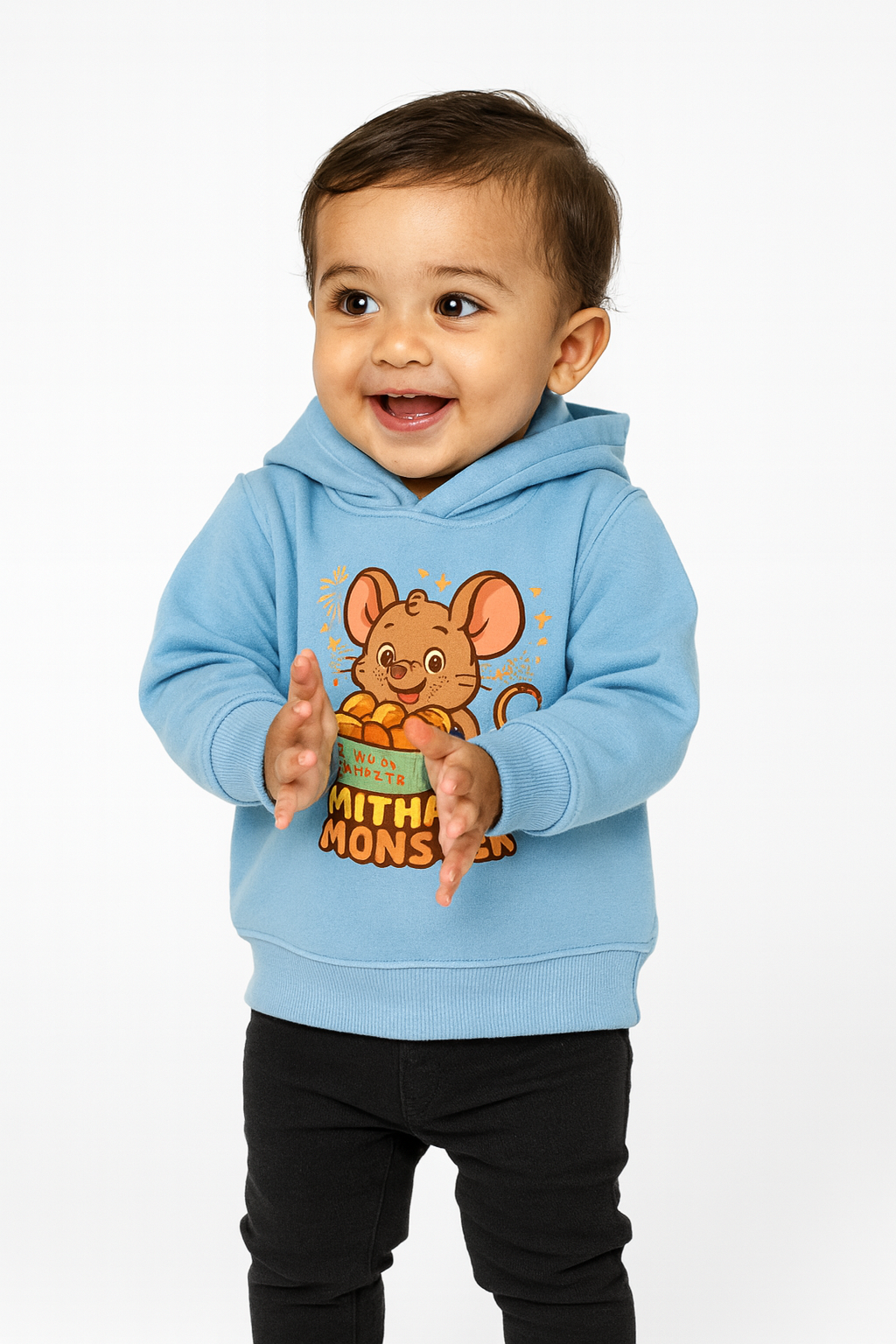 Smiling toddler wearing Mithai Monster mouse hoodie holding laddoos on sky blue fabric.